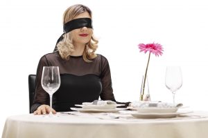 Young woman with blindfold on her eyes sitting at a romantic dinner table isolated on white background